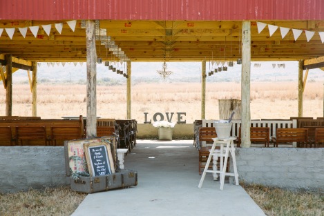 Open air chapel with LOVE word and candle chandelier, white ladder holding a bucket of confetti and a vintage trunk with signage